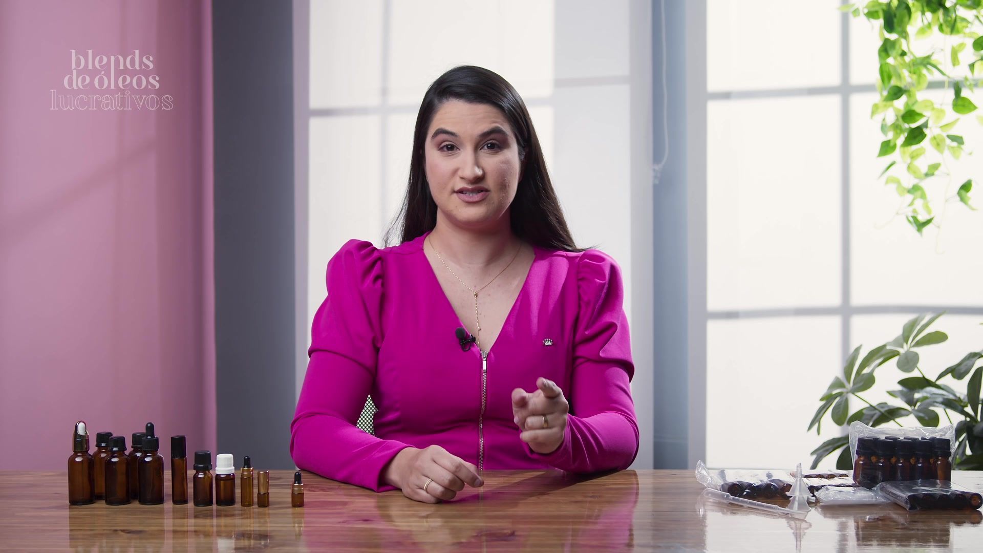 Uma mulher de camisa rosa está sentada a uma mesa com frascos de óleos essenciais.
