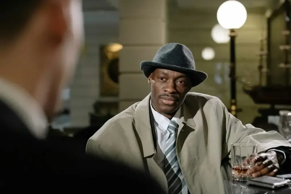Man in a fedora and trench coat seated at a table, speaking in a dimly lit room