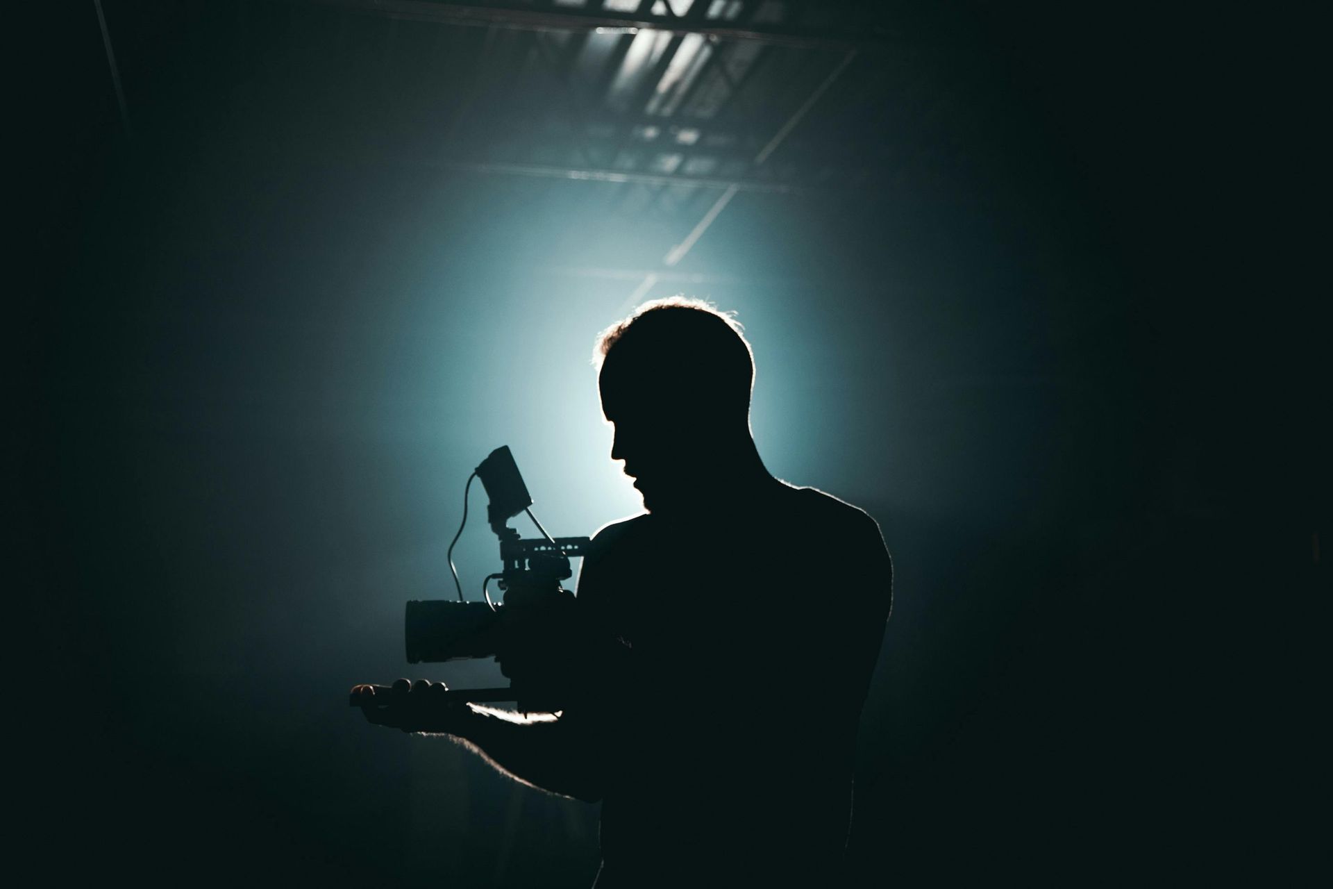 Silhouette of a person holding a video camera against a bright light in a dark space.