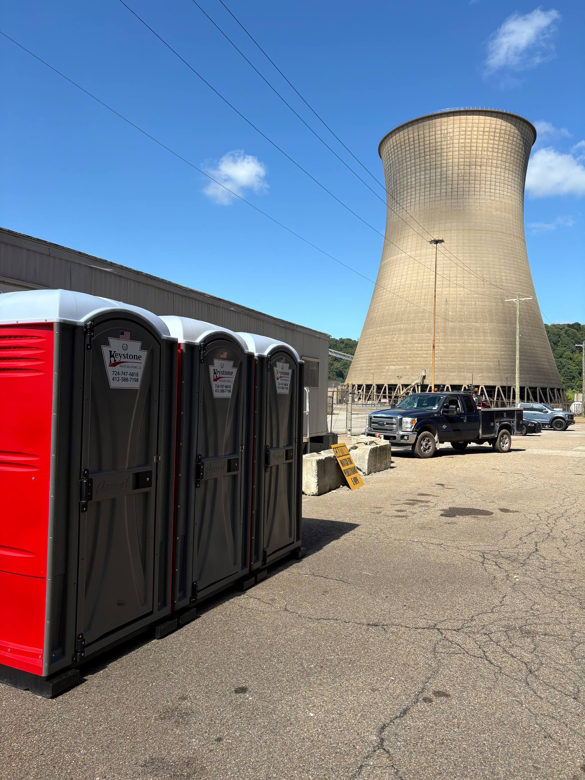 Row of grey portable toilets with red sides on a paved area, trees in the background. | Keystone Waste Solutions