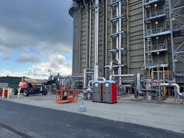 A construction site with two red portable toilets, a parked boom lift, and industrial pipes against a tall factory wall.