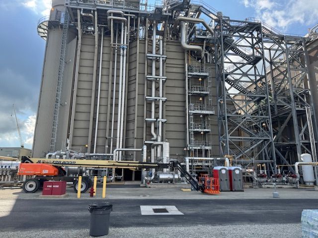 A tall industrial facility with large pipes and metal scaffolding, featuring a construction lift parked in the foreground.