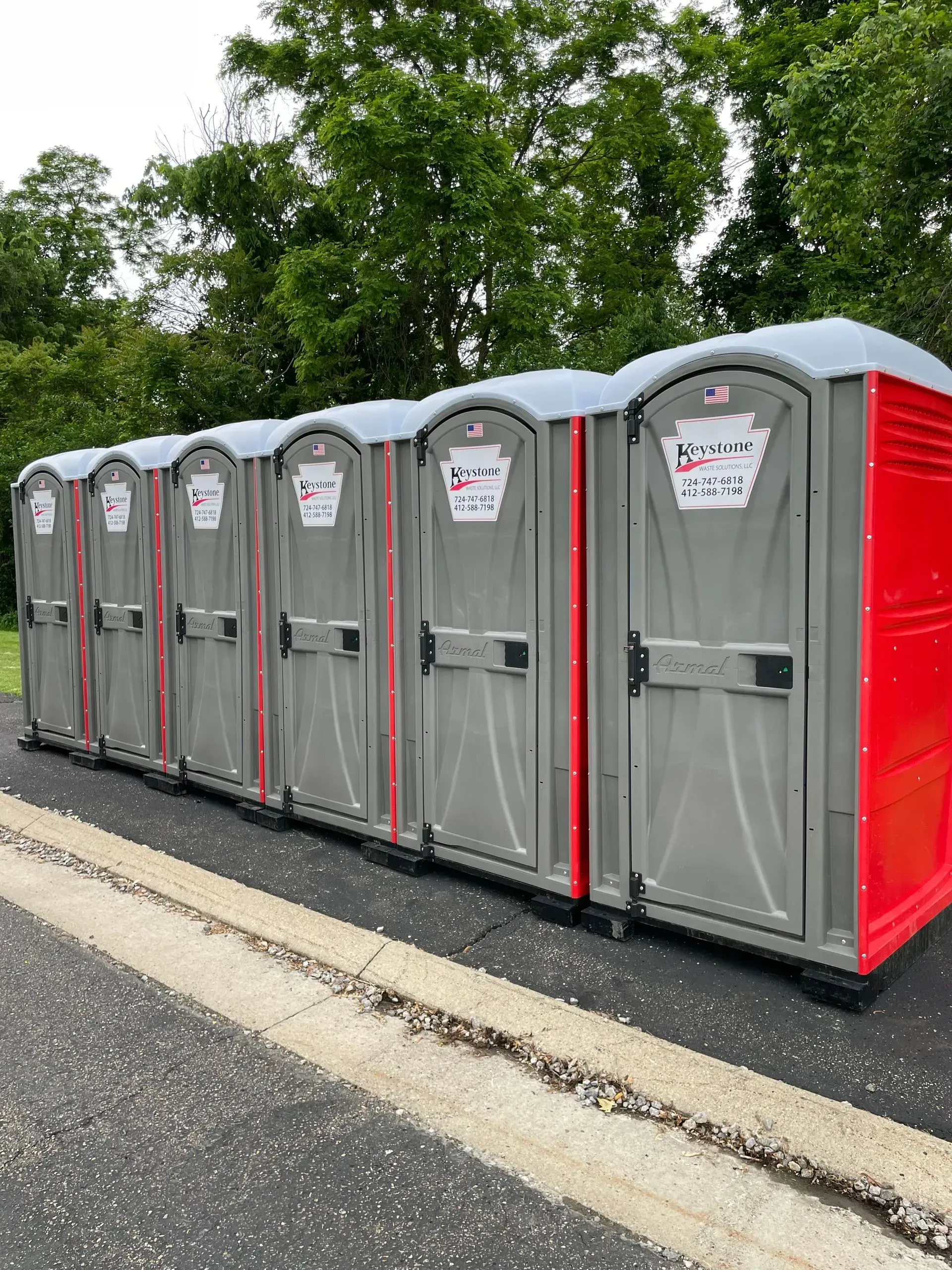 A row of six gray and red portable toilets positioned outdoors on an asphalt surface with green trees in the background.