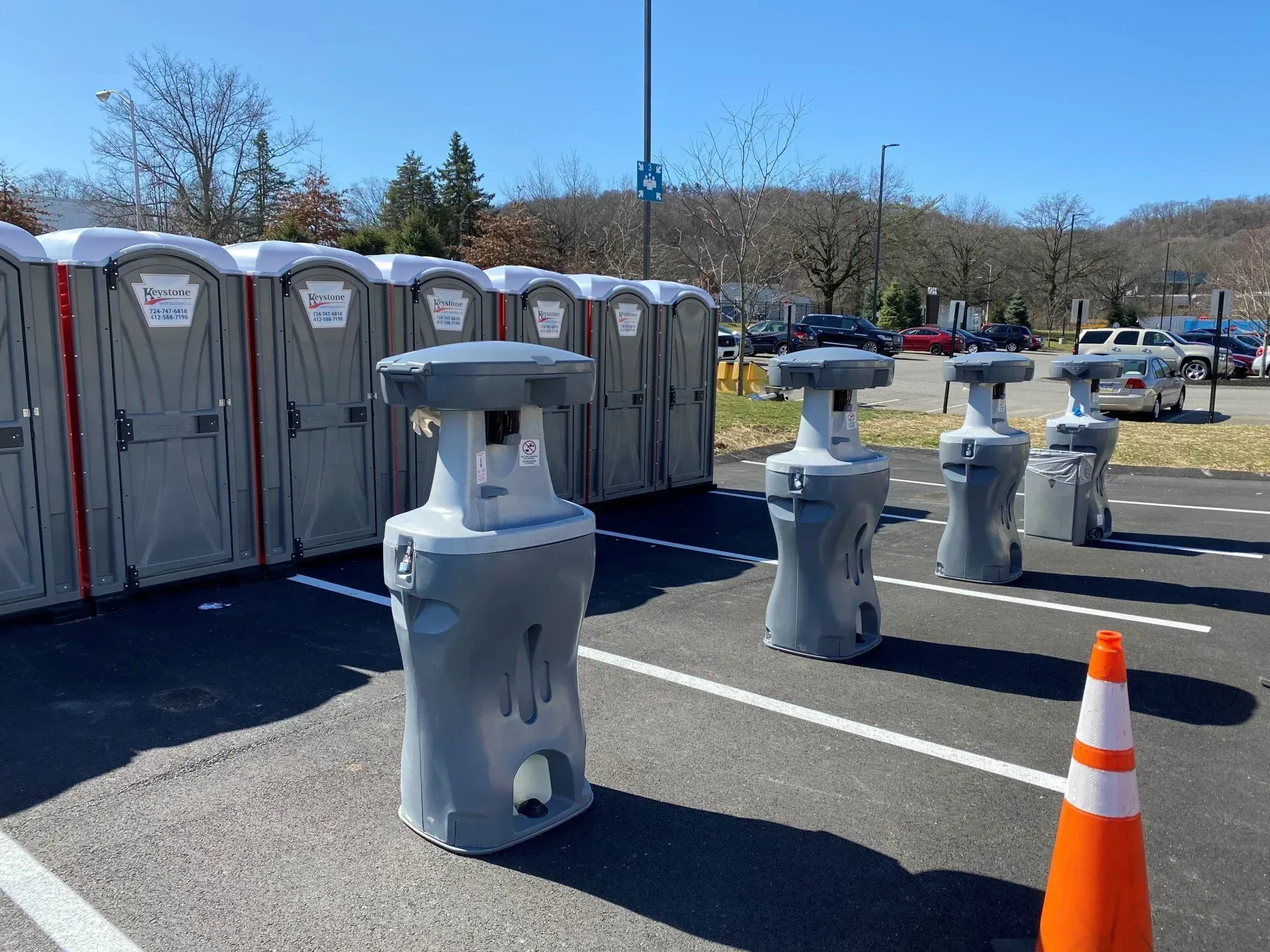 A row of portable toilets and several gray freestanding handwashing stations set up in a sunny outdoor parking lot.