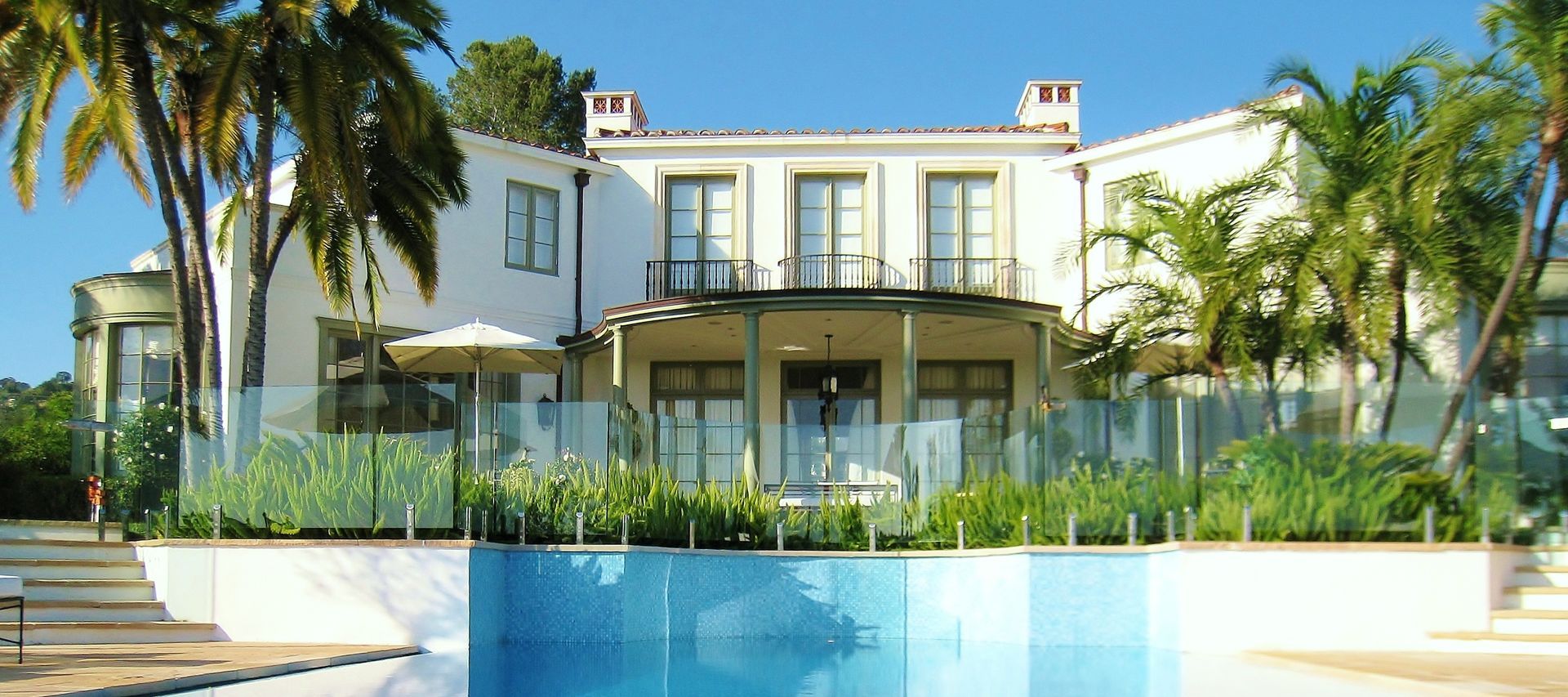 Large white building with a pool in front, surrounded by palm trees and glass fencing.