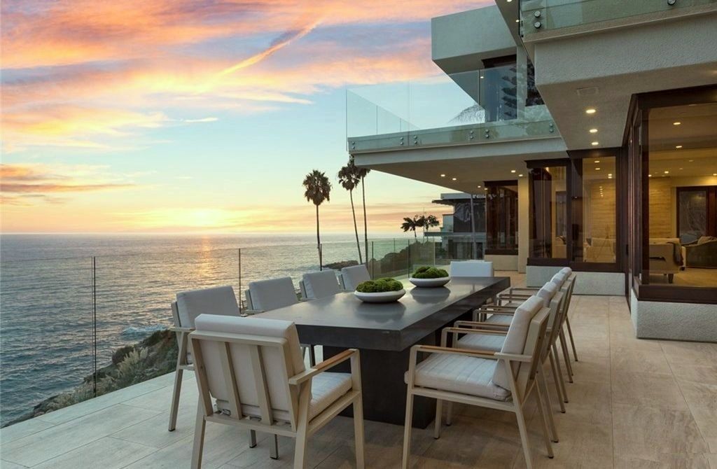 Outdoor dining area overlooking the ocean at sunset. Table with chairs, palm trees, and modern house.