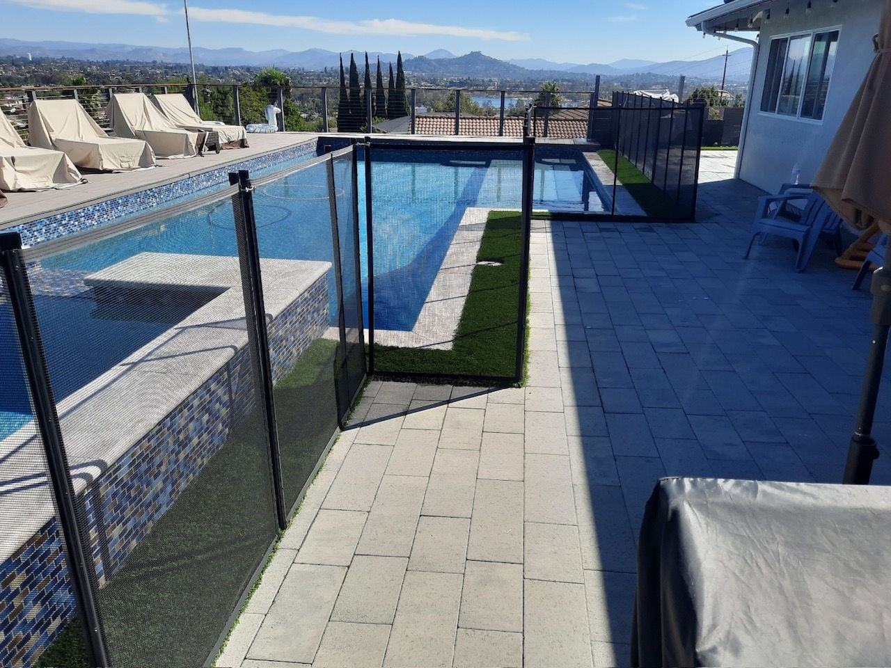 A pool with a black safety fence, patio, and a view of distant hills.