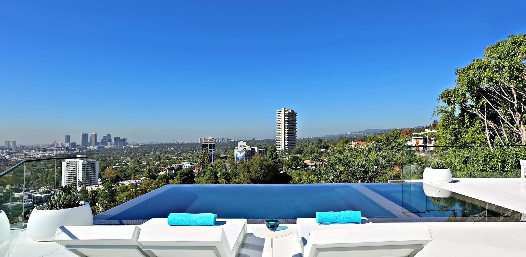 Pool overlooking a cityscape, with blue water, white lounge chairs, and a clear sky.