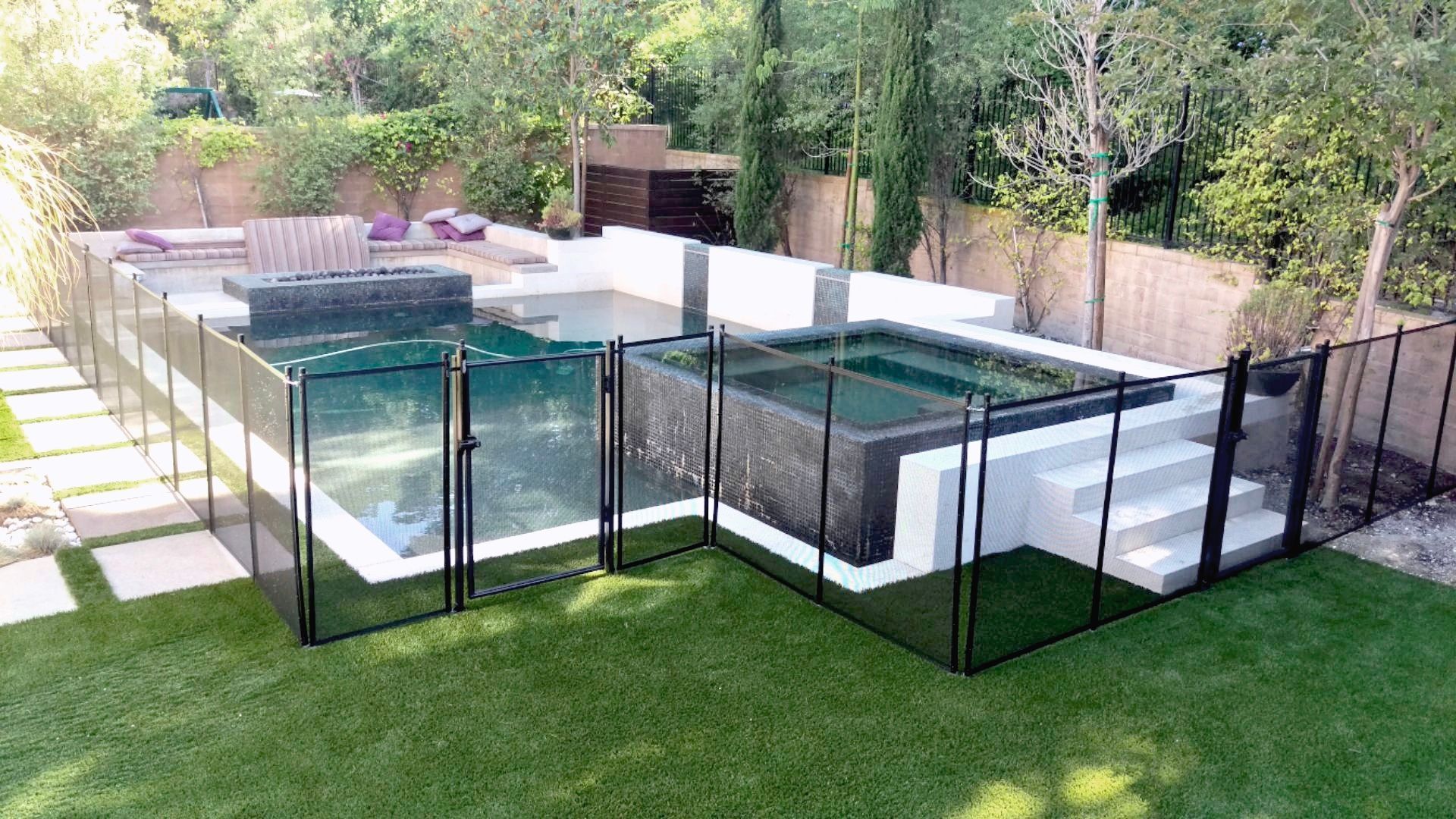 Backyard with pools and spas, fenced with black mesh, on green lawn. Concrete steps and patio furniture are present.