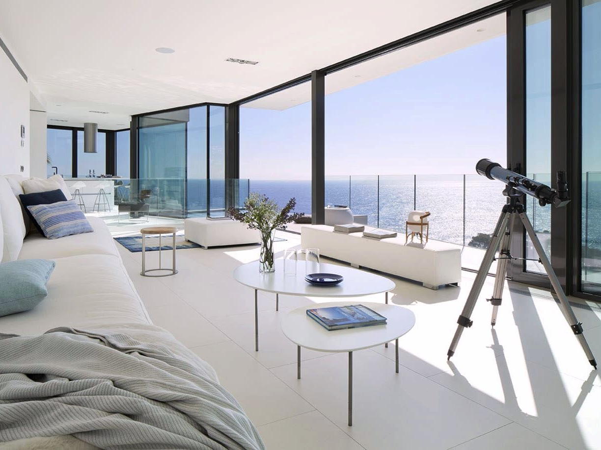 Modern living room with white decor, floor-to-ceiling windows, and ocean view. Telescope on tripod.