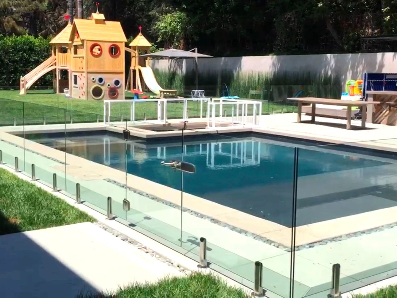 Backyard pool with glass fence, small play structure, and green lawn.