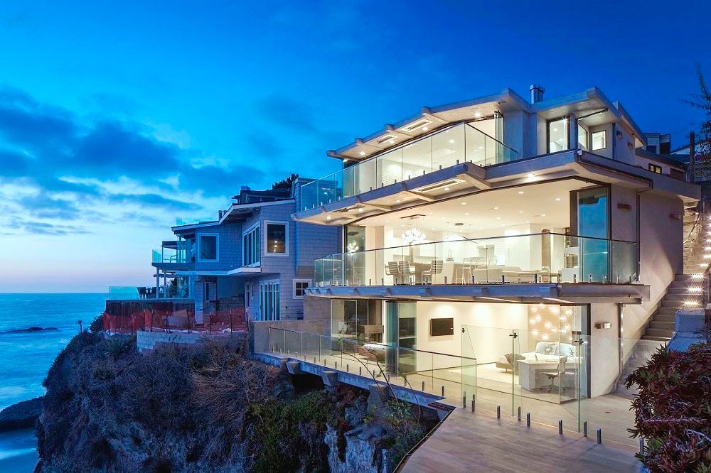 Luxury coastal home with glass walls, overlooking the ocean at dusk.