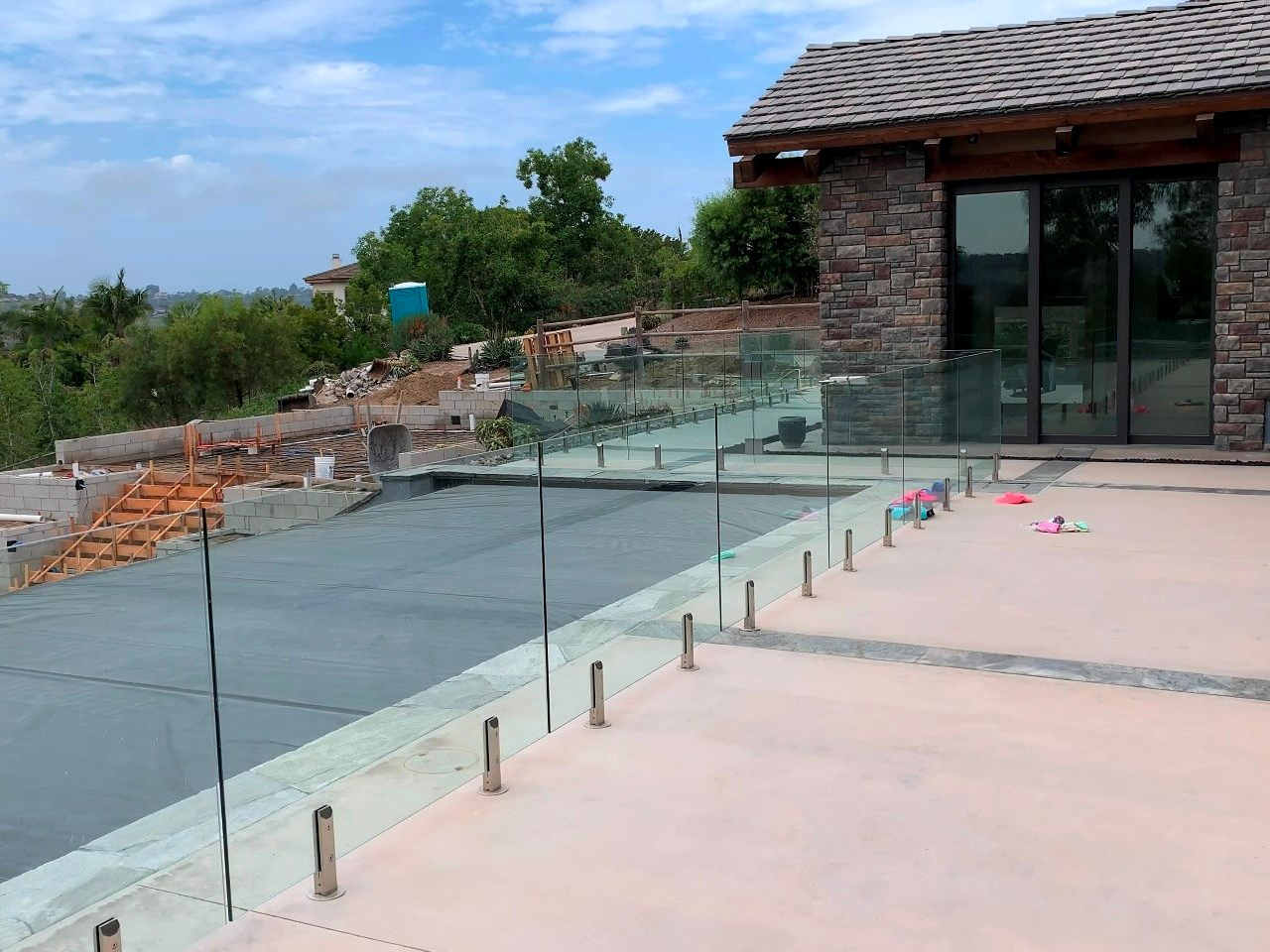 Glass railing around a concrete patio with a house in the background. Blue sky and trees visible.