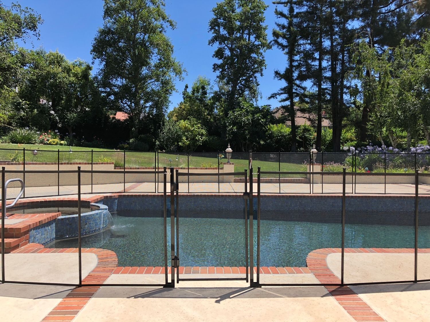 Pool surrounded by a dark mesh safety fence with a gate. Brick deck, blue water, and trees in the background.
