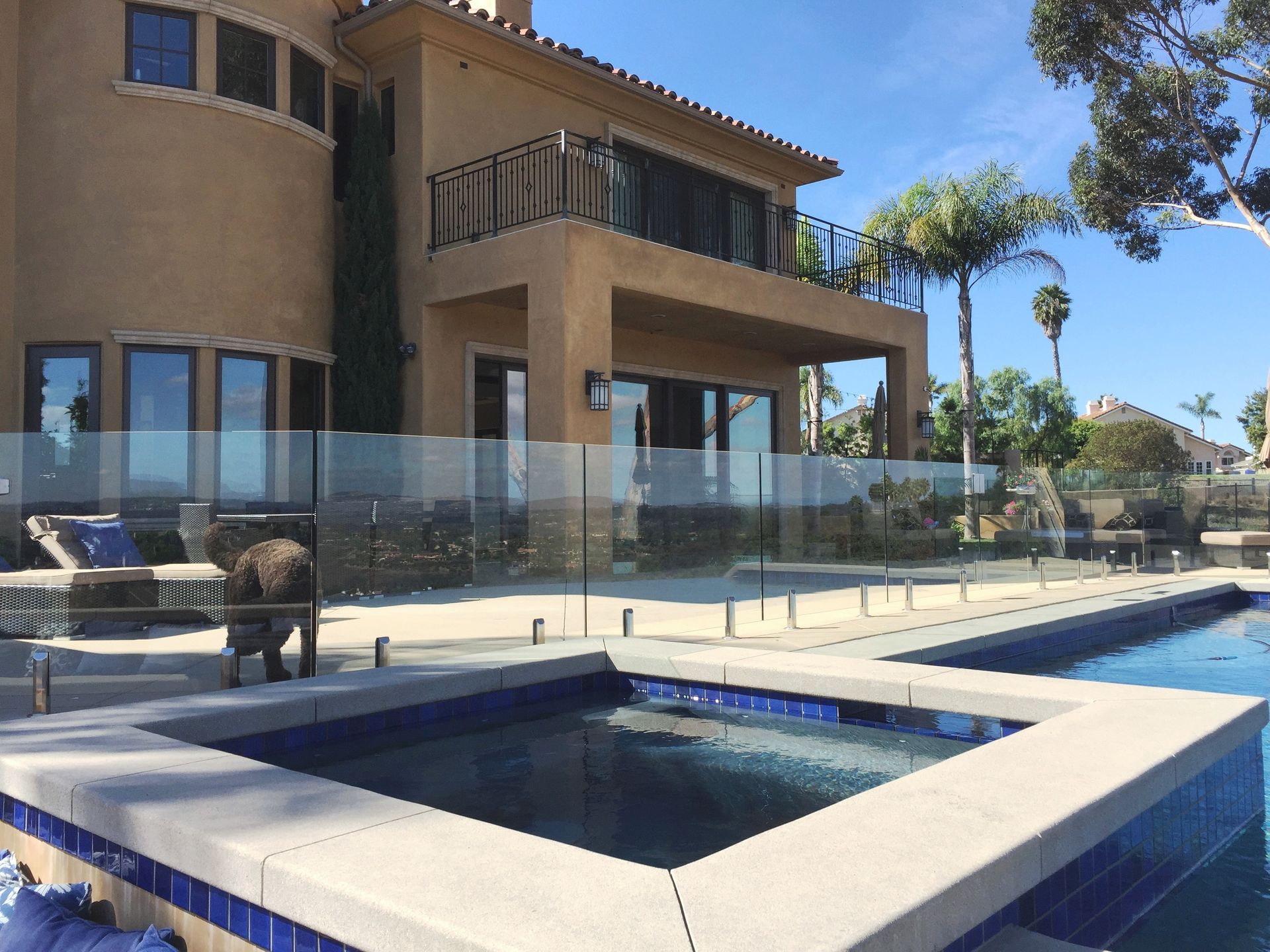 A modern house with a pool and spa, featuring a glass fence and a dog. Sunny day, blue water.
