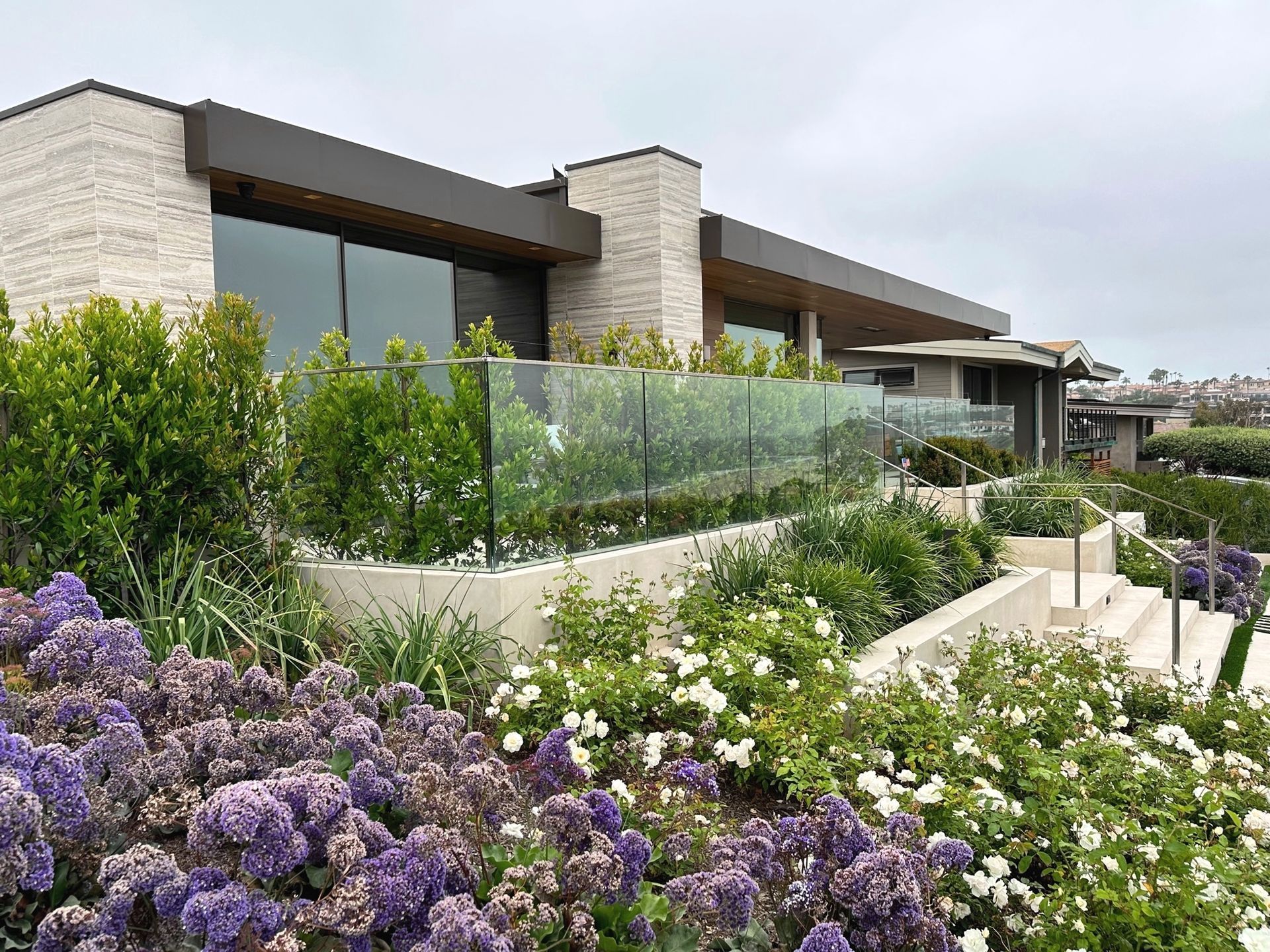 Modern house with glass railing, tiered landscaping, and flowering bushes under an overcast sky.