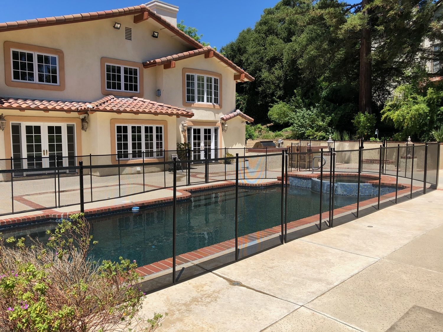 Backyard pool with black mesh fence, next to a two-story beige house with a red tile roof.