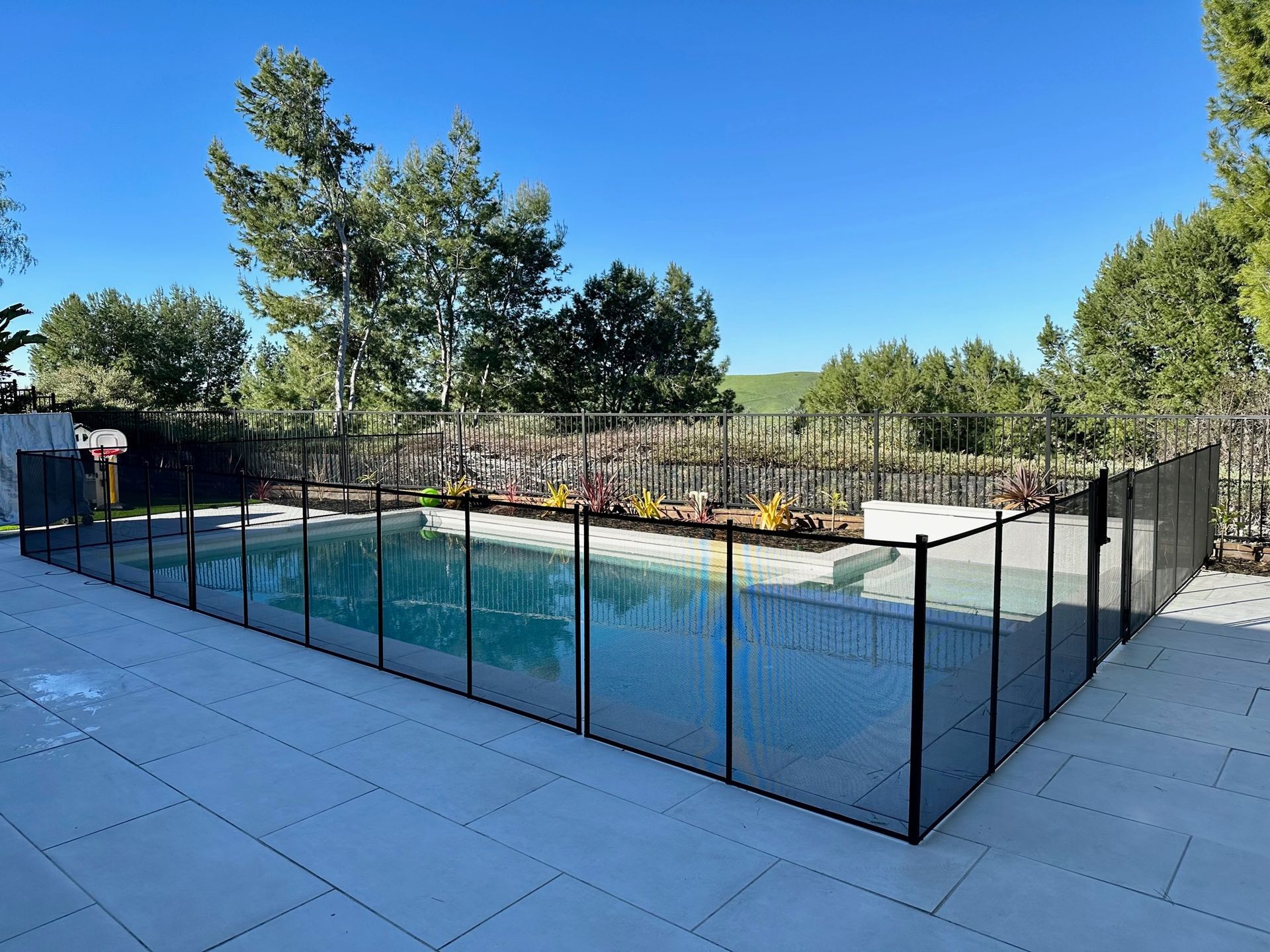 Black mesh safety fence surrounds a rectangular swimming pool on a gray patio. Trees in background.