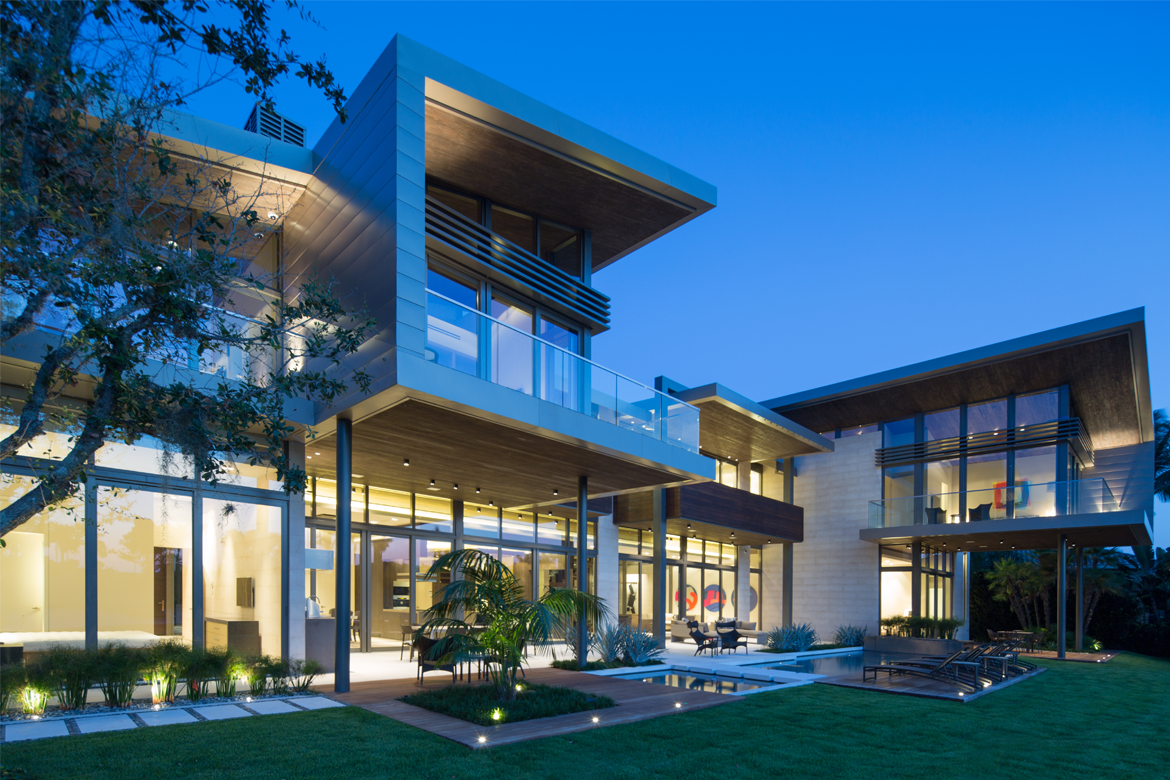 Modern two-story house with large windows and overhanging roof lit at dusk. Landscaped yard, blue sky.