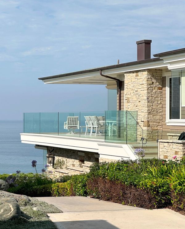 Oceanfront house with a glass-railed balcony, stone exterior, and partial ocean view.