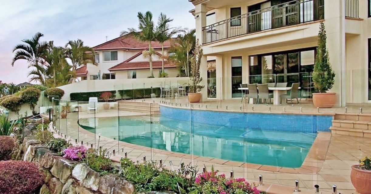Luxury home with pool; blue water, stone retaining wall, patio, palm trees, and cloudy sky.