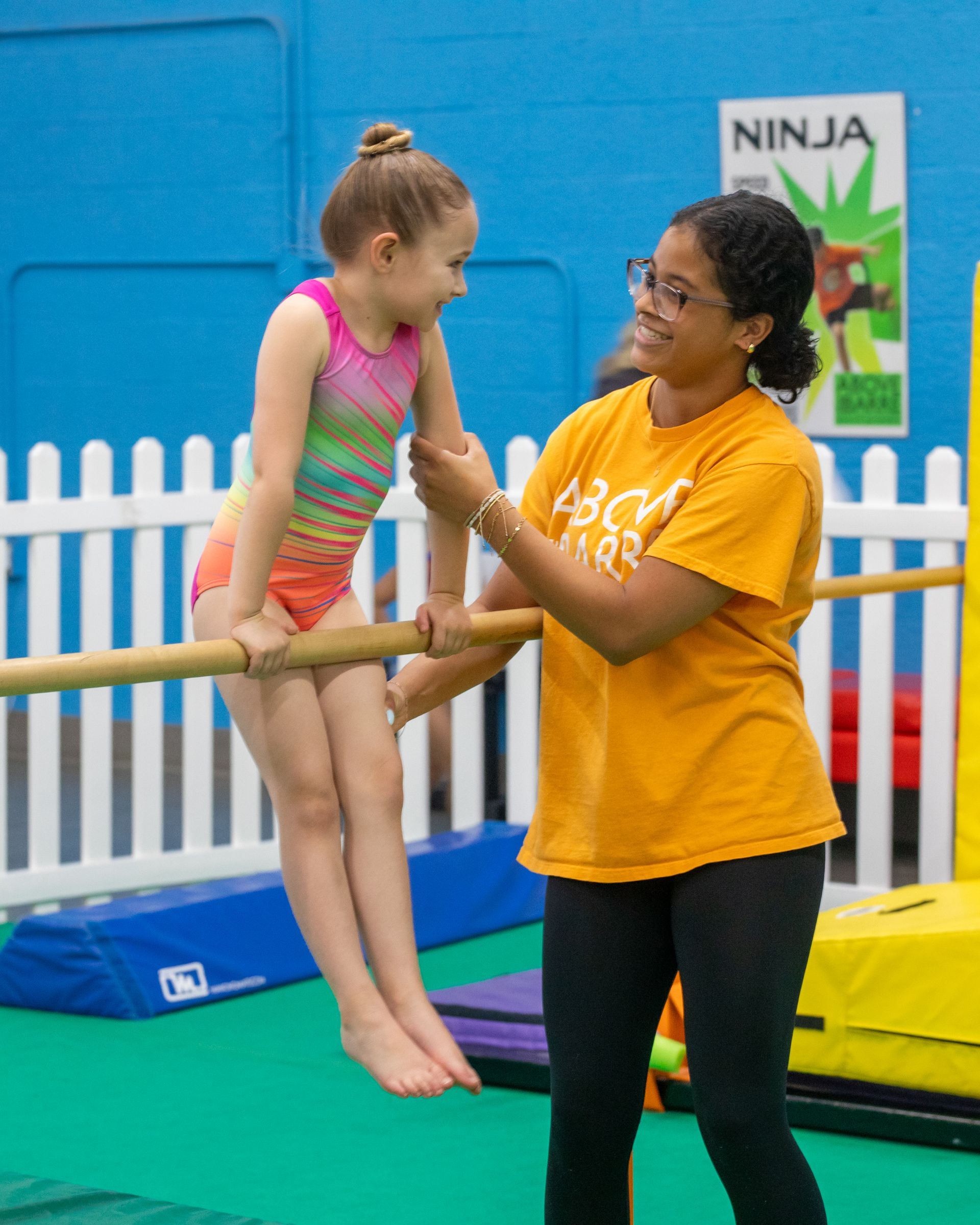 Boy and girl sit on floor, instructor facing them. Gymnastics gym setting, green floor, mats, colorful equipment.