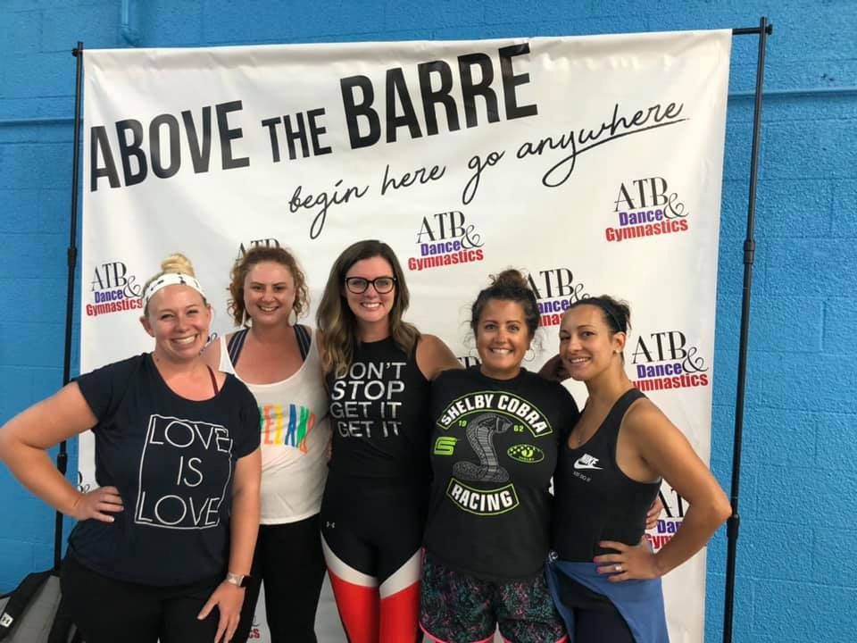 Five women pose in front of a banner. The banner reads 