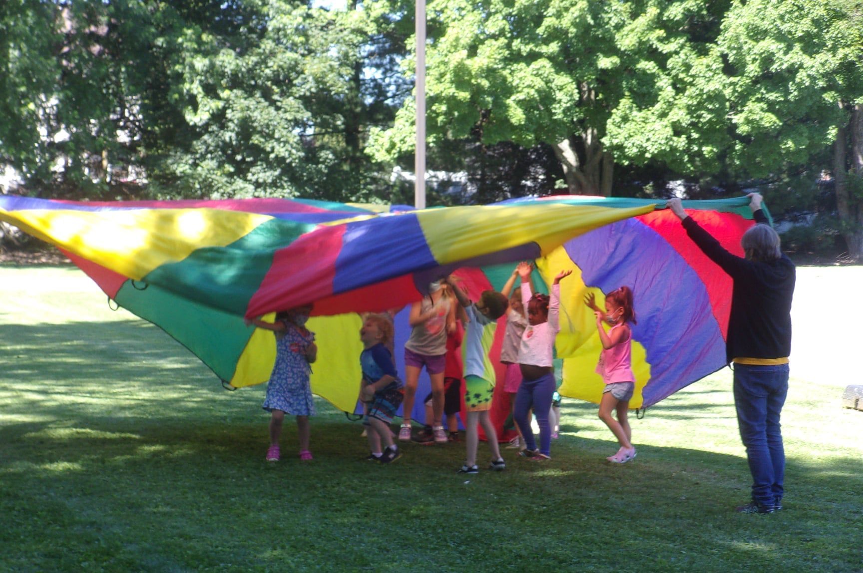 A group of people standing in a circle outdoors, holding up a large, colorful, circular parachute above their heads.