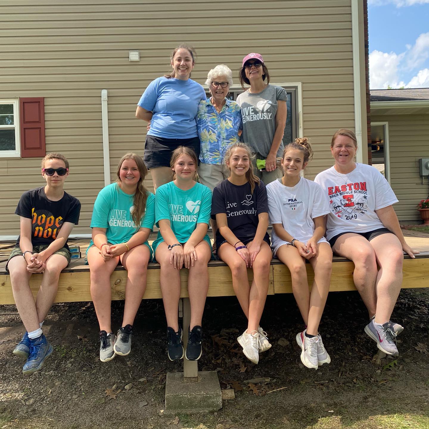 A group of ten people stand and sit on a wooden deck in front of a tan house, smiling at the camera.