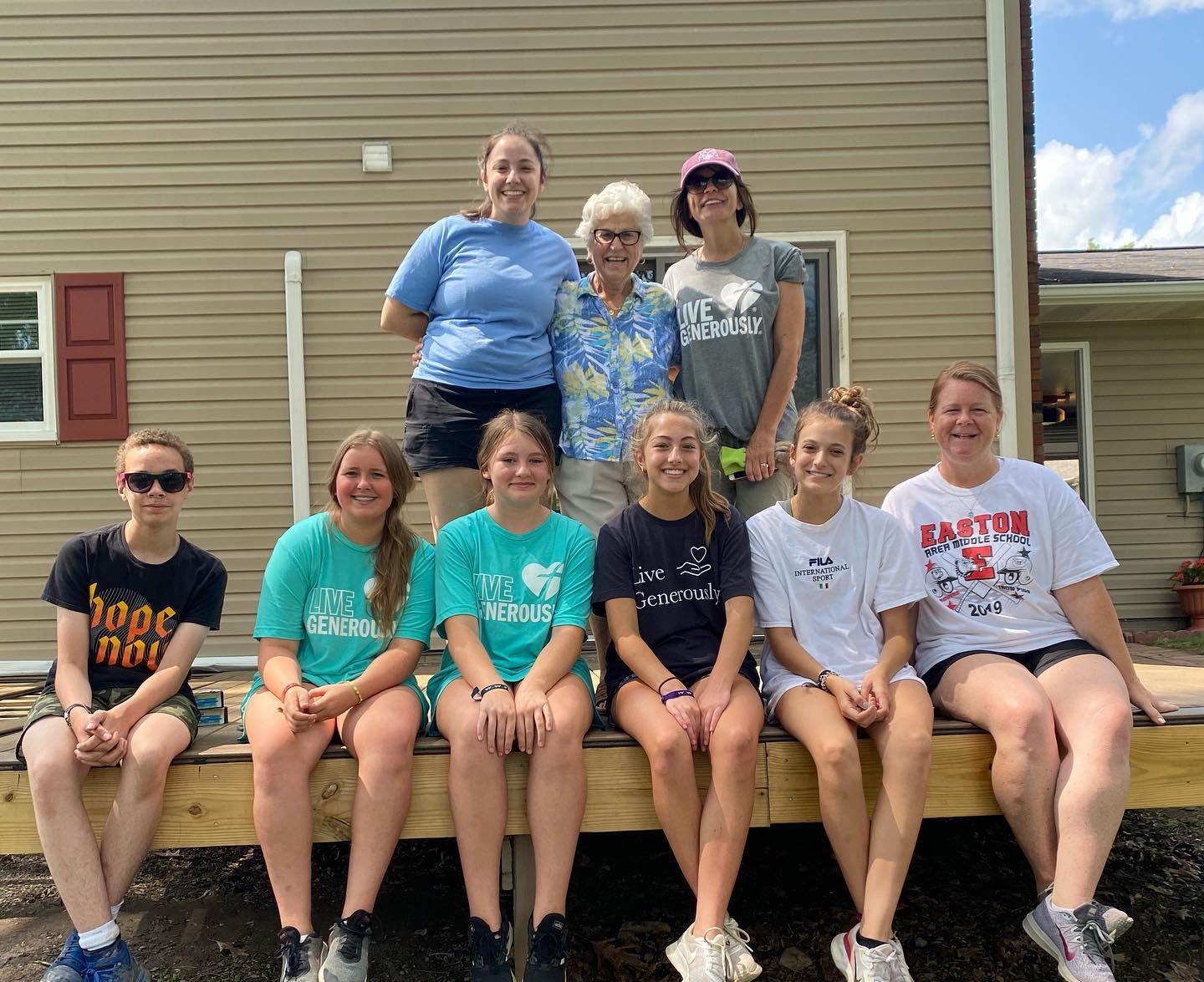 A group of ten people stand and sit on a wooden deck in front of a house with tan siding, smiling for a group photo.