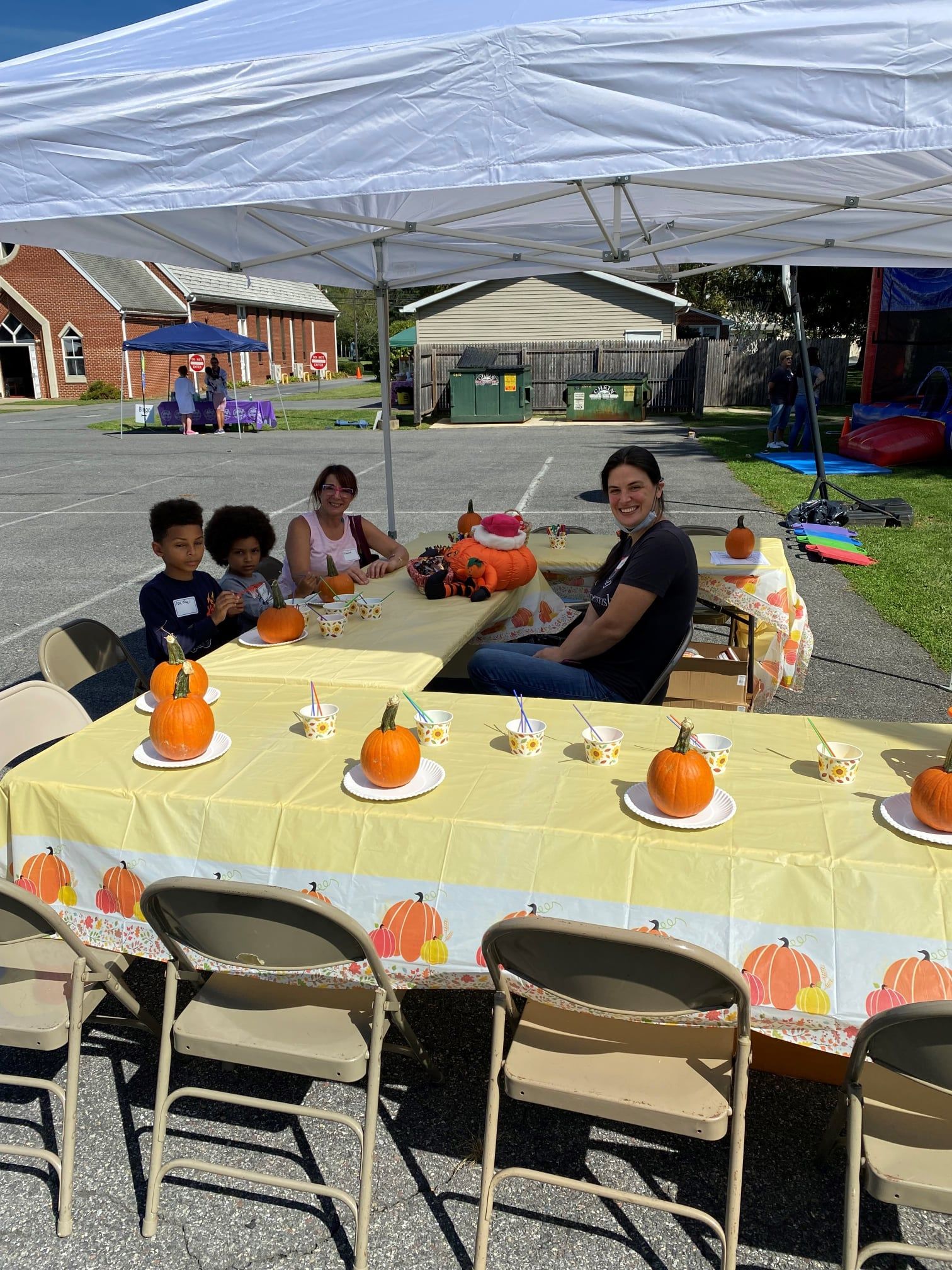 People seated at a table under a white tent with small pumpkins, preparing for a festive outdoor community event.