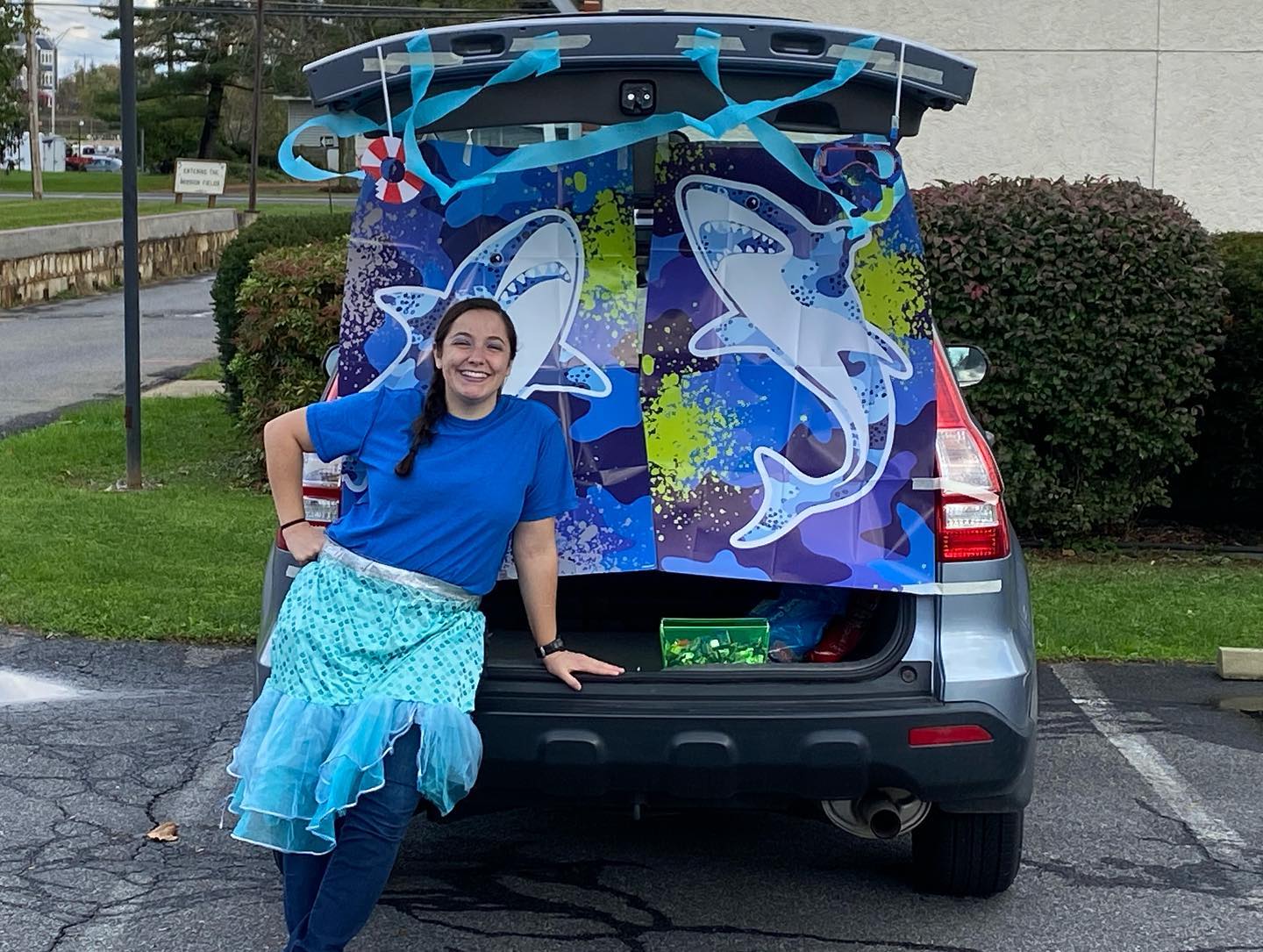 A person in a mermaid-style skirt poses by a car trunk decorated with a shark-themed backdrop for a trunk-or-treat event.