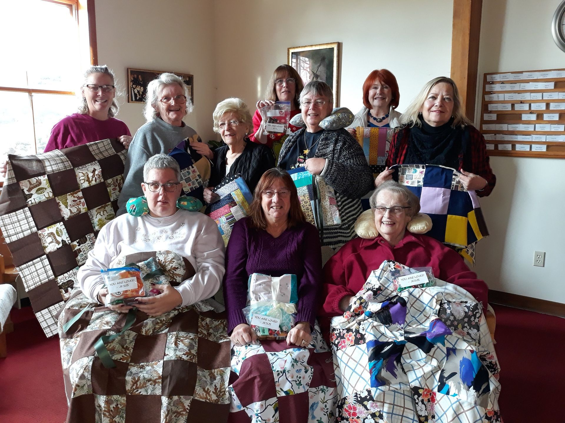 A group of people holding homemade quilts in a room with a bulletin board and a red carpet.
