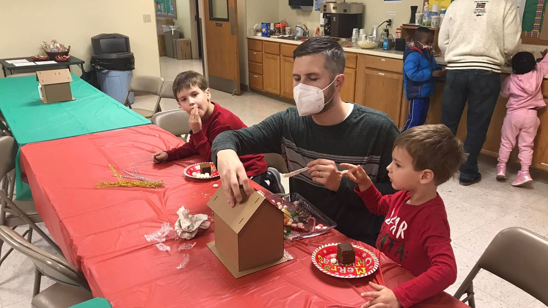 A man and two children sit at a red-covered table in a classroom decorating cardboard houses.