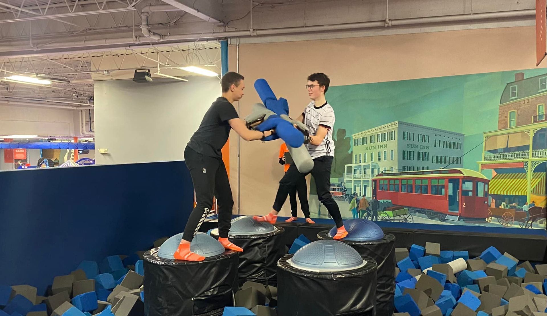 Two people stand on balancing pedestals in a foam pit, engaging in a friendly foam-padded jousting match.