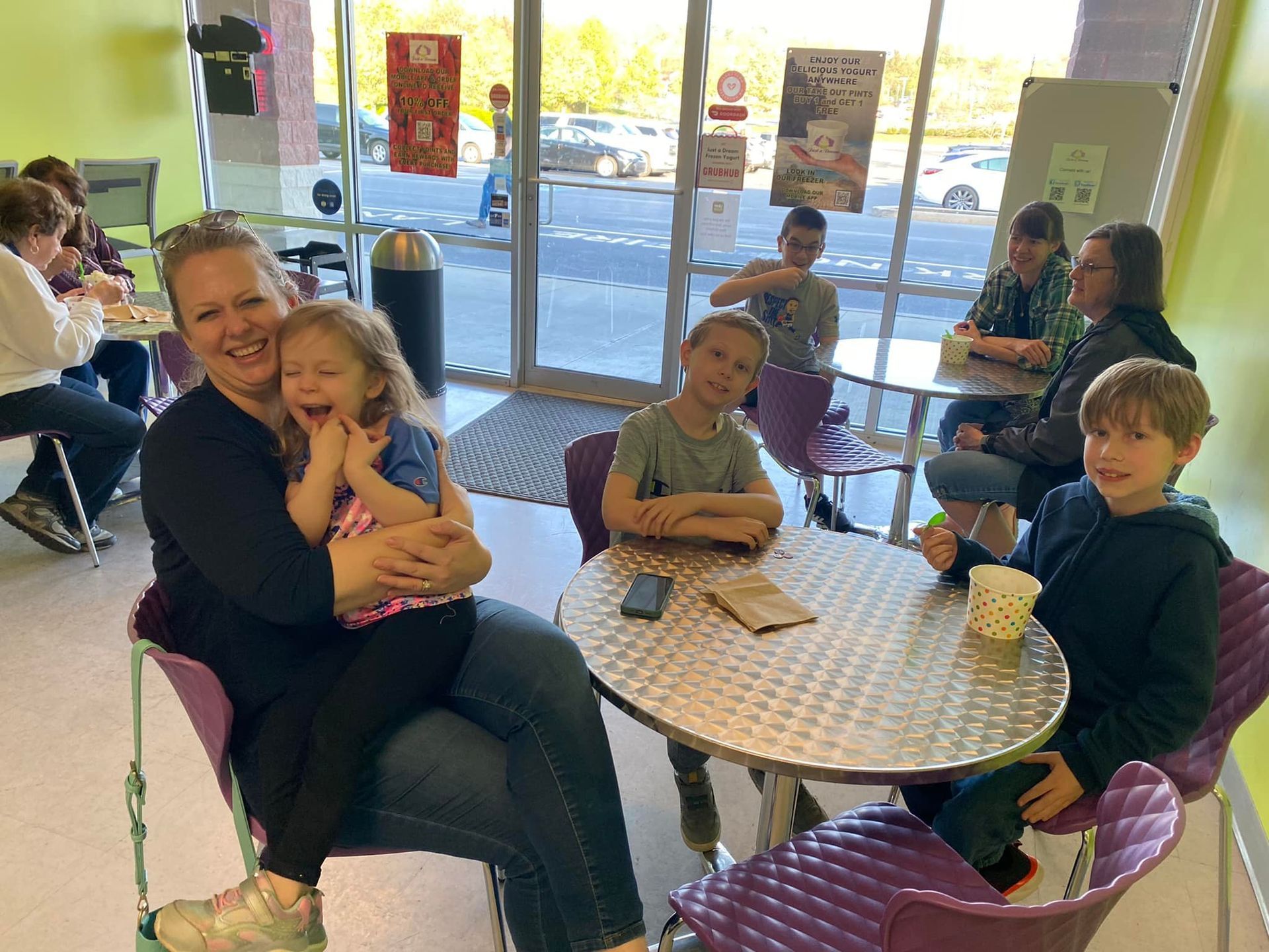 A woman holds a small child on her lap in a room with people sitting at tables, with large windows in the background.