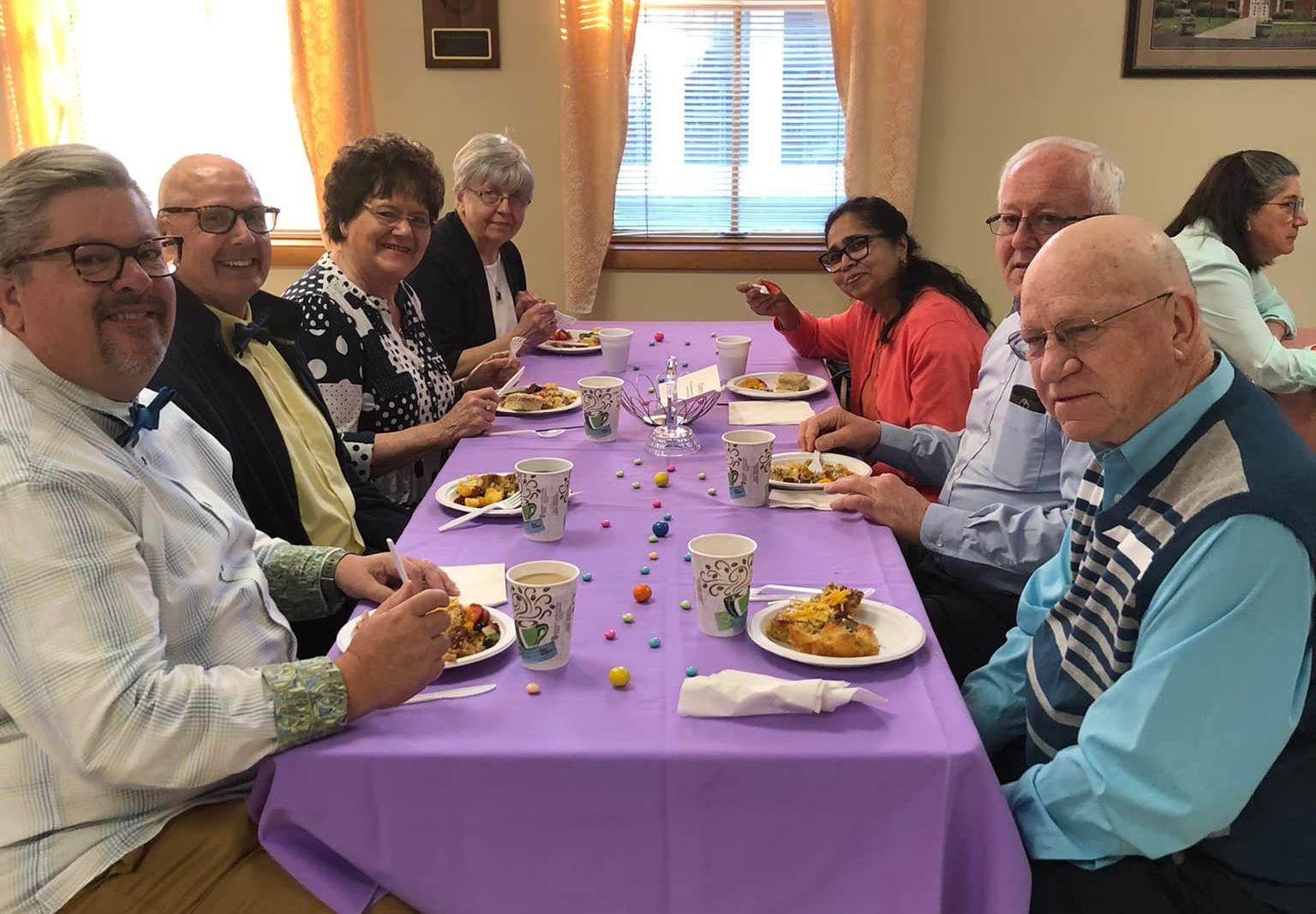 A group of people are eating and socializing around a long table covered with a purple cloth in a brightly lit room.