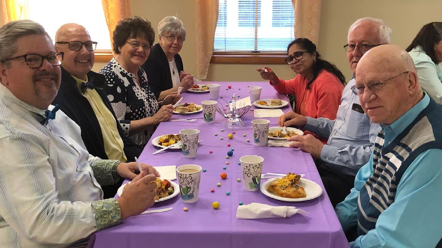 Seven people sit at a rectangular table with a purple cloth, eating a meal in a well-lit dining area.