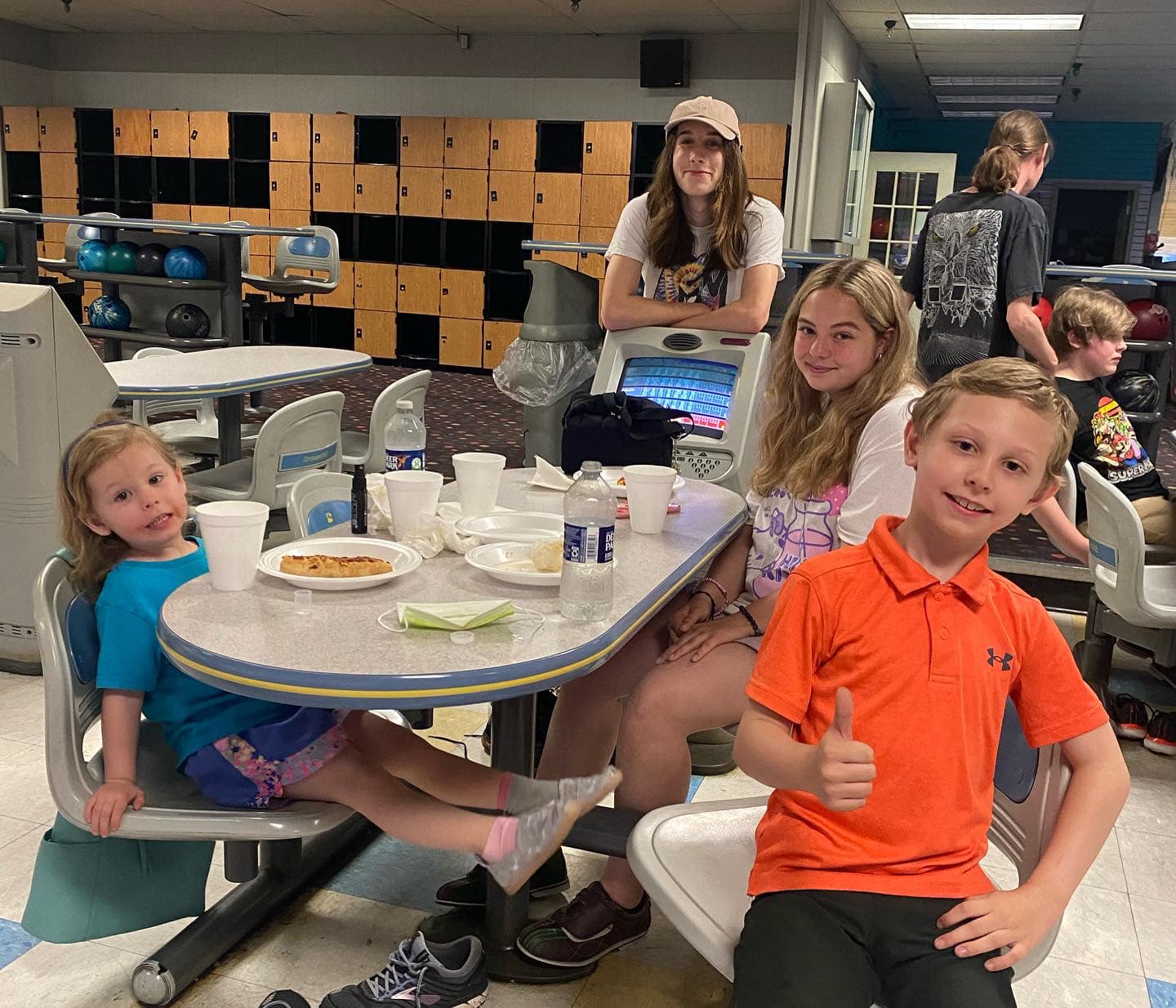 Four people sitting at a table in a bowling alley, with a child in an orange shirt giving a thumbs-up in the foreground.