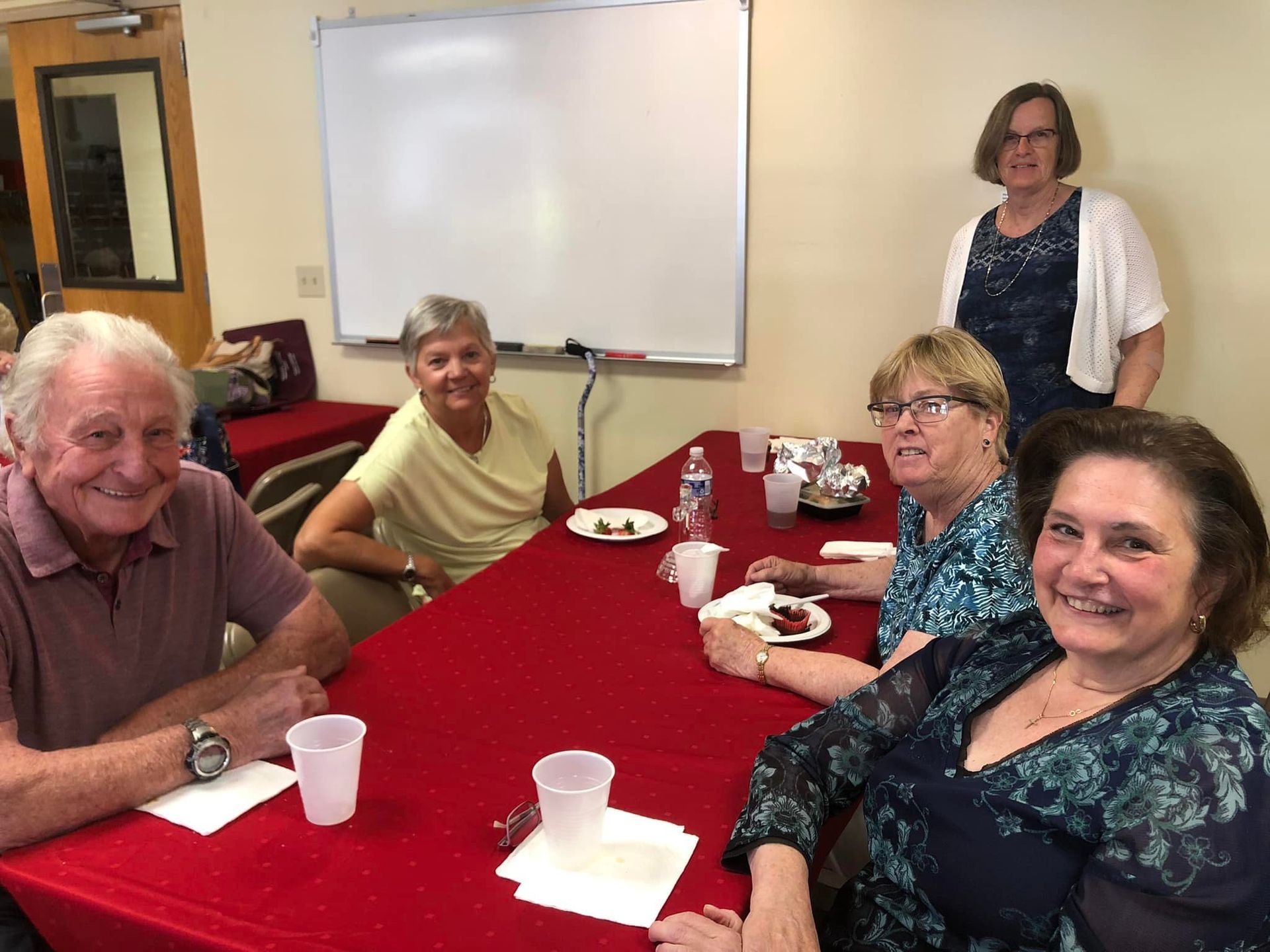 A group of five people smile while seated around a table covered in a red tablecloth in a room with a whiteboard.