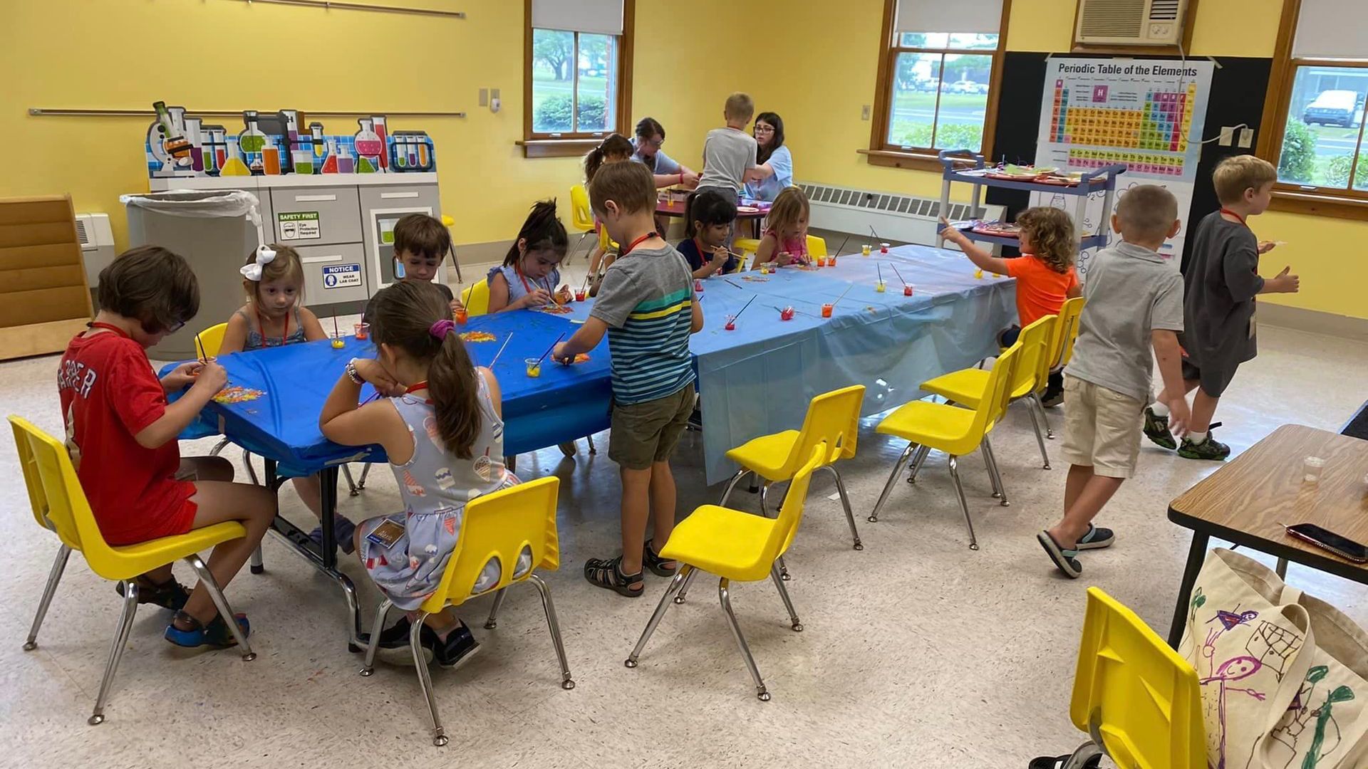 Children work at a long blue-covered table in a bright classroom with yellow walls and chairs.