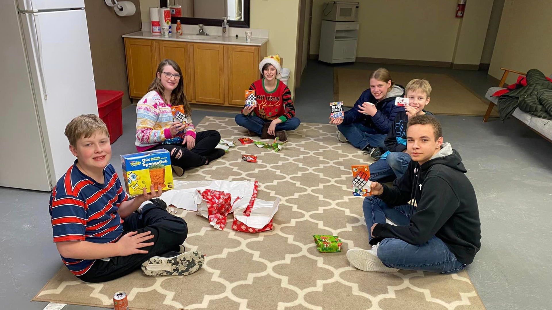 Seven people sit on a patterned rug in a room, opening gifts and smiling at the camera.