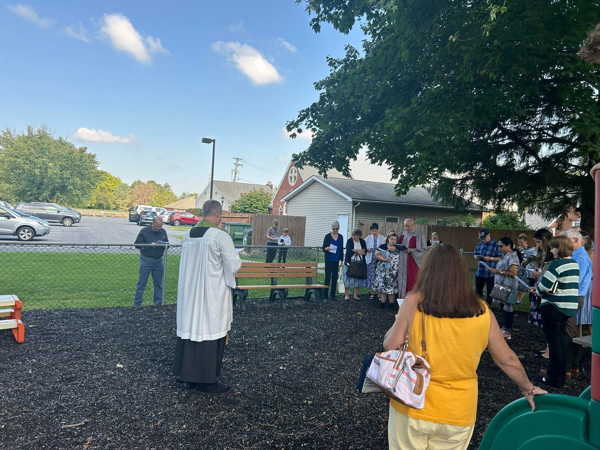 A person in liturgical vestments stands facing a group of people gathered outdoors on a wood-chipped surface near a fence.