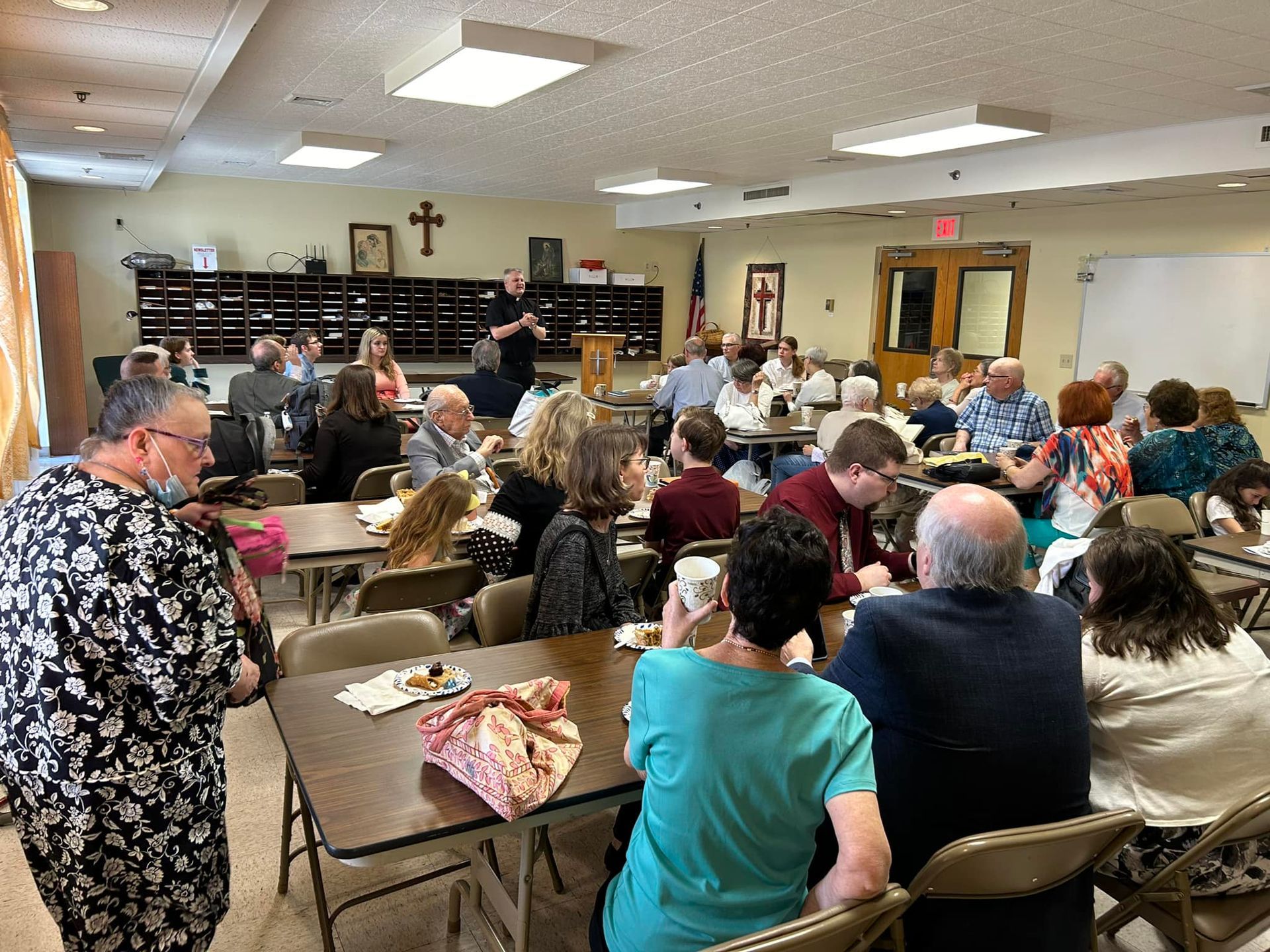 A group of people seated at tables in a brightly lit community hall, attending an indoor gathering or meeting.