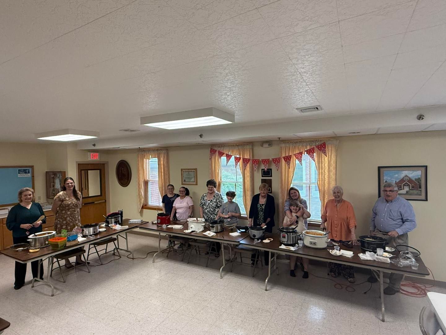 A group of people standing behind a long table set with food in a community room decorated with a banner.