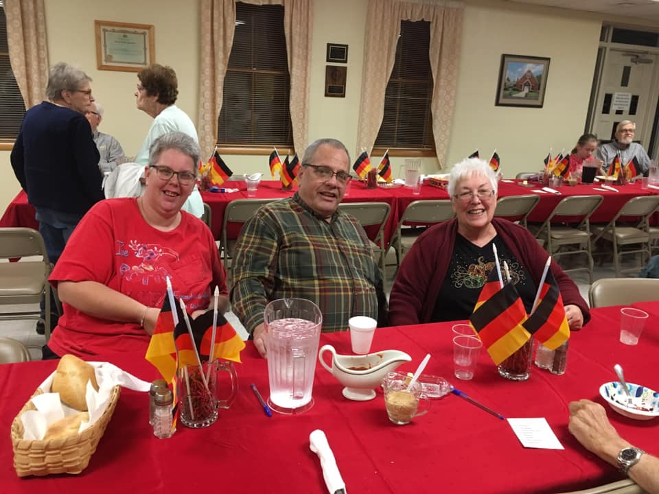 Three people sit at a table covered with a red tablecloth and German flag decorations, posing for a photo.