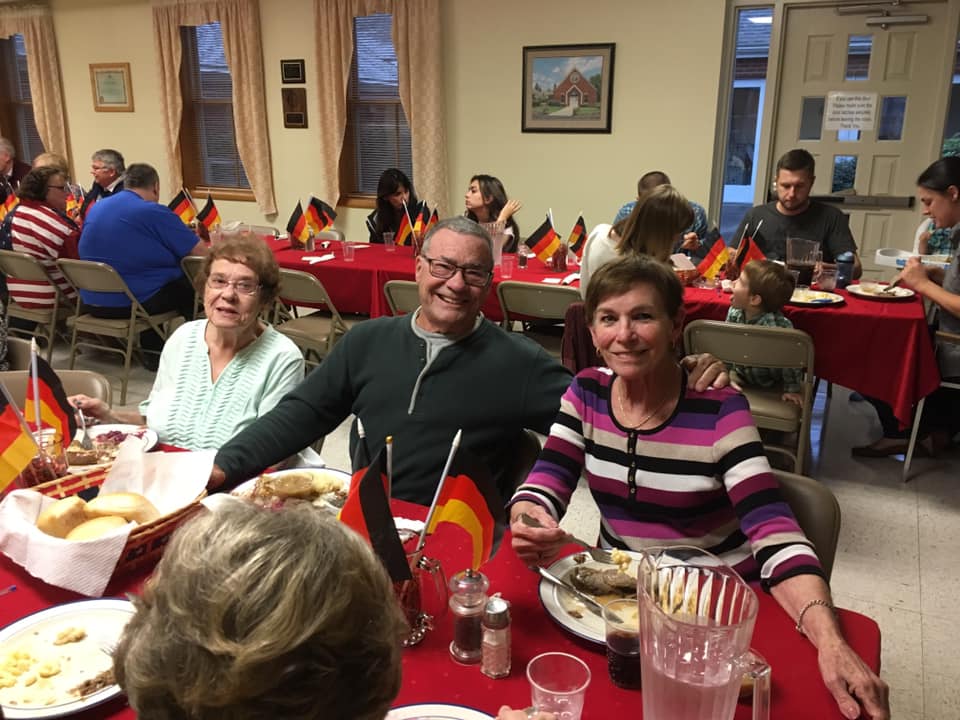 People seated at round tables covered in red tablecloths decorated with small German flags at a community event.