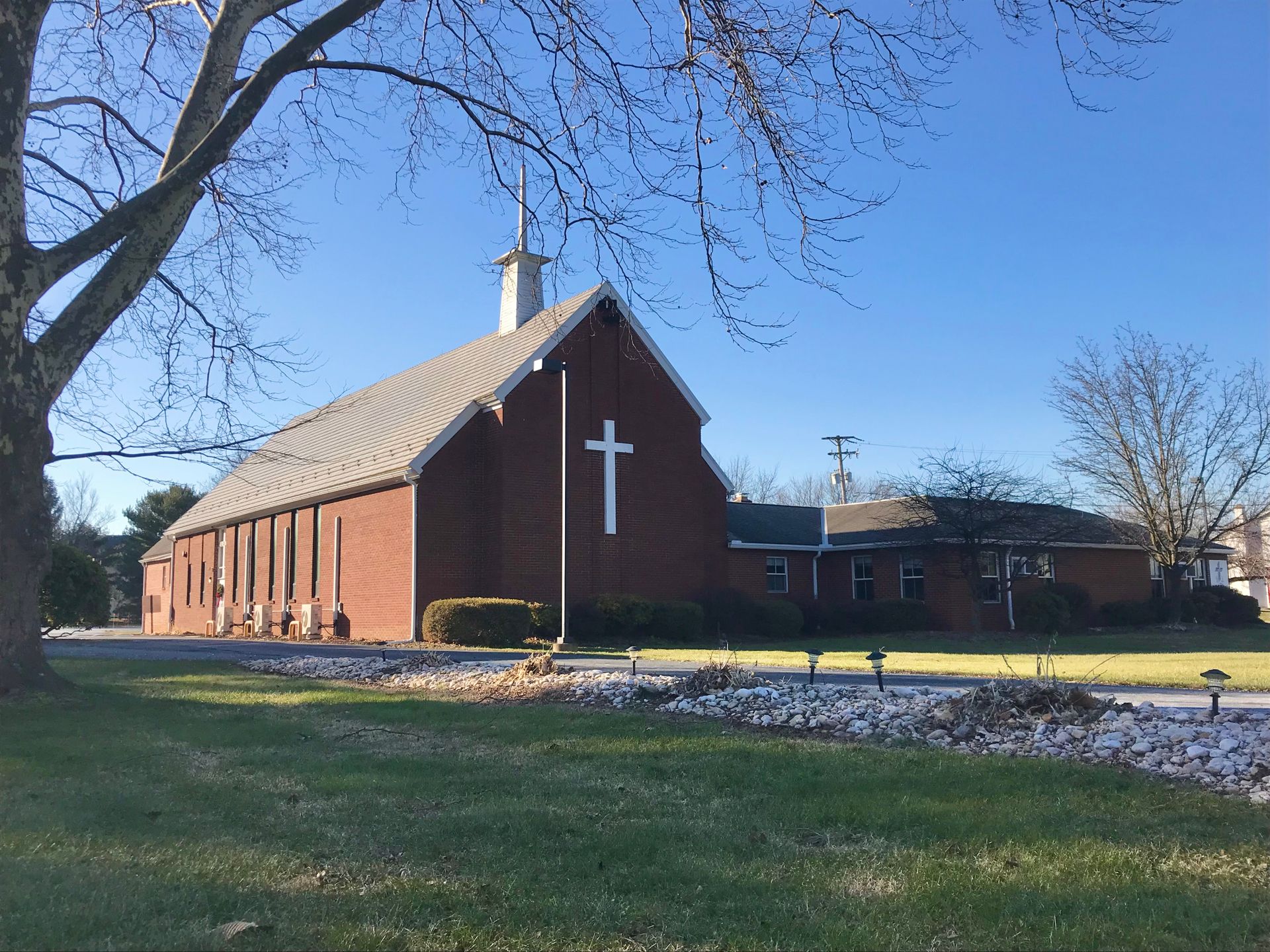 A red brick church with a white cross on its side stands on a sunny, grassy lawn under a clear blue sky.