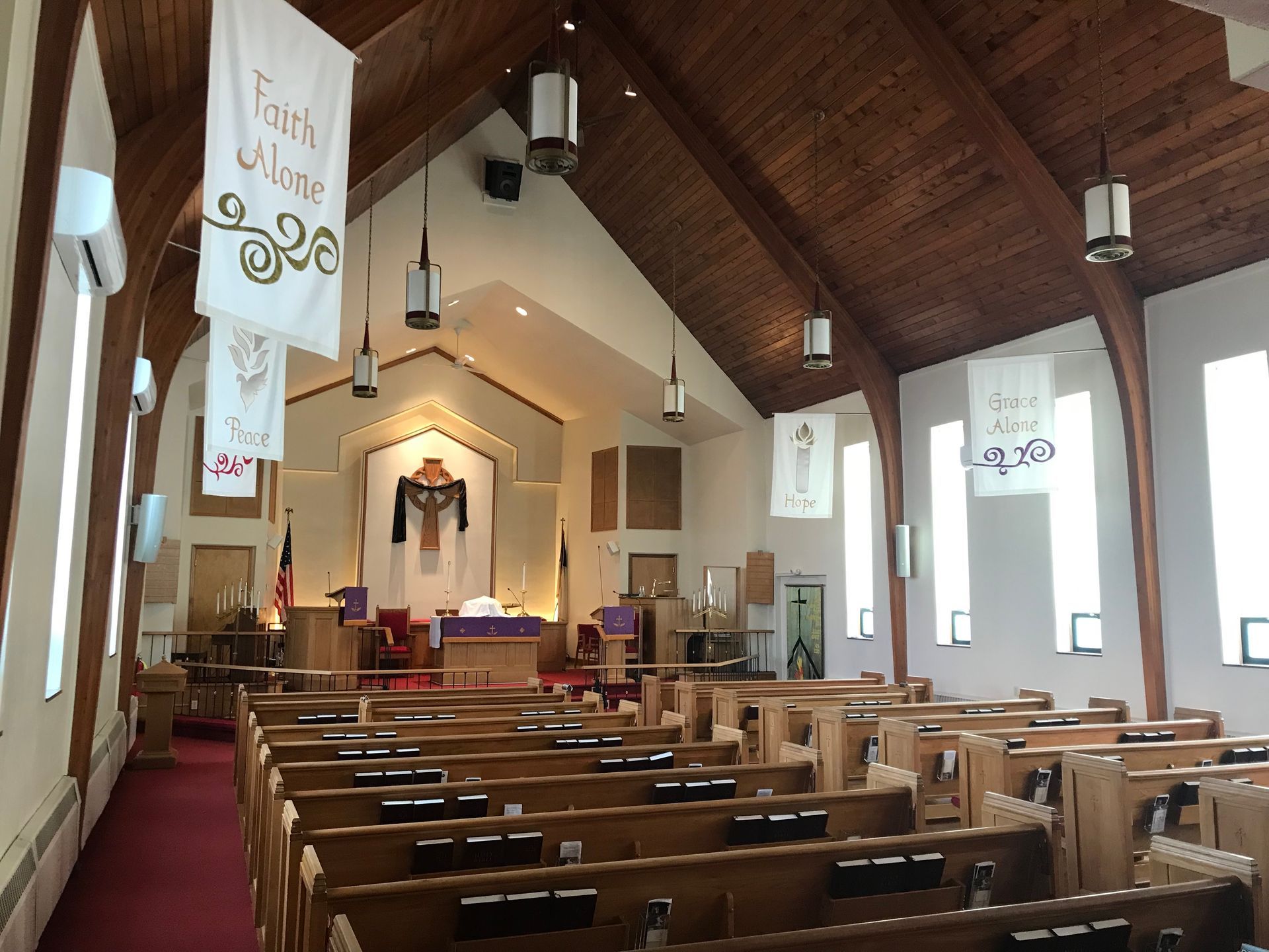 Interior of a church with wooden vaulted ceilings, rows of pews, a red carpet runner, and white hanging banners.