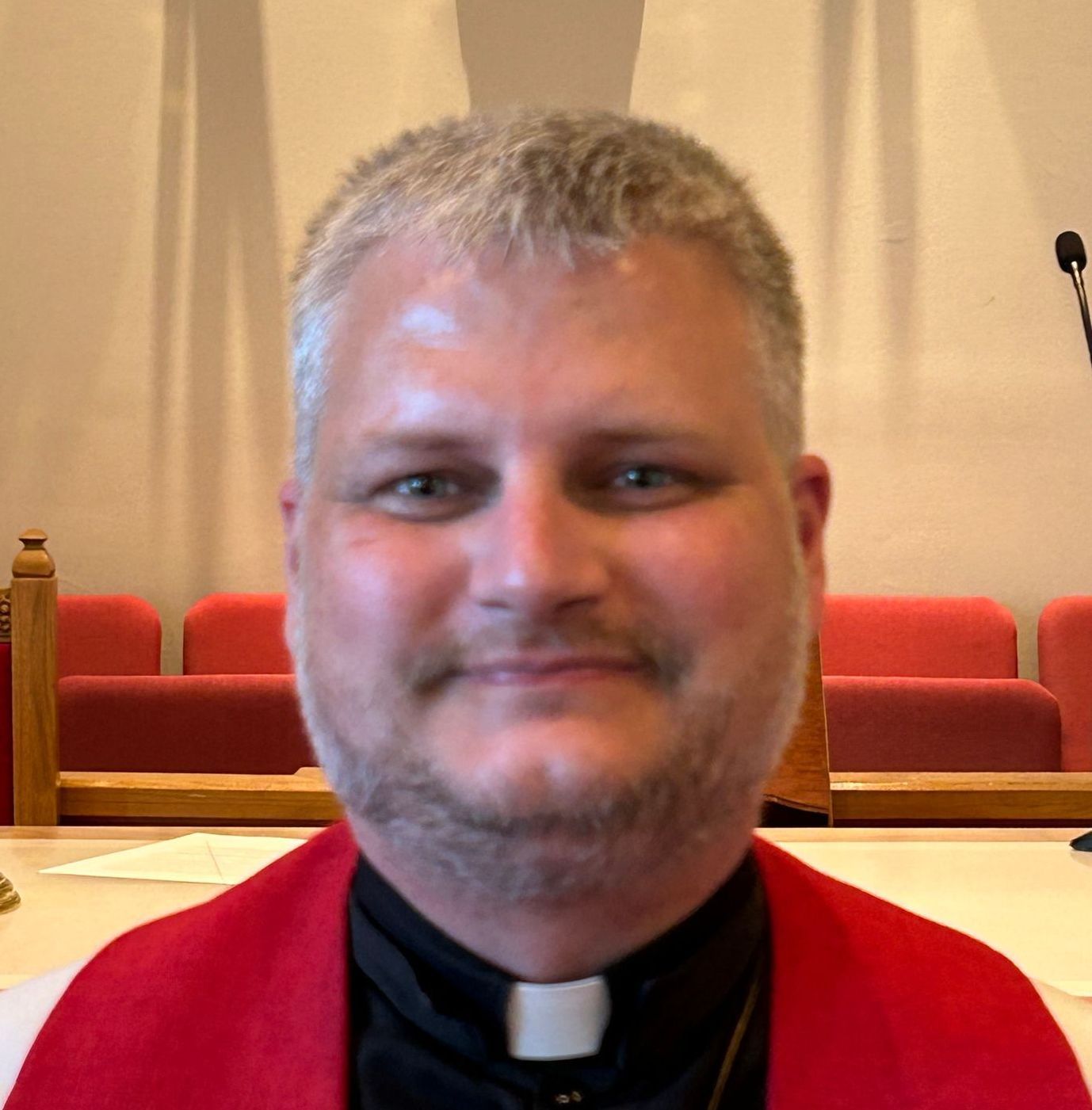 A person wearing a clerical collar and a red stole, standing in a church with red seating in the background.